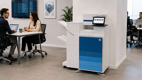 Ein moderner HP-Drucker mit Touchscreen steht in einem Büro in der Nähe einer Wand. Im Hintergrund sitzen zwei Personen an einem Tisch und unterhalten sich; im Hintergrund sind Computerbildschirme und die Büroeinrichtung zu sehen.