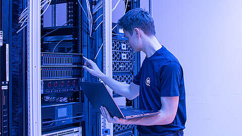 Q-FOX® Mitarbeiter drückt auf einen Kopf an einem Serverschrank und hat einen Laptop in der Hand