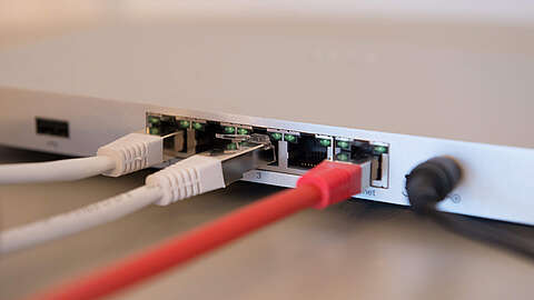 Nahaufnahme der Rückseite eines Netzwerk-Switches mit mehreren Ethernet-Kabeln - weiß, grau und rot -, die in die Anschlüsse eingesteckt sind, und einem schwarzen Stromkabel, das auf der rechten Seite angeschlossen ist.