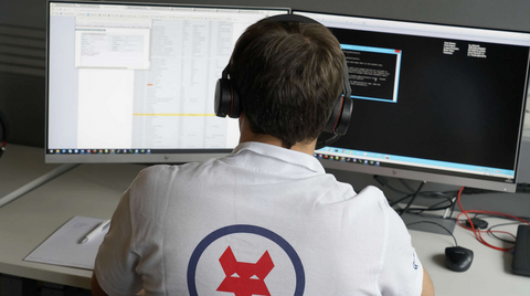 Eine Person, die Kopfhörer und ein weißes Hemd mit einem rot-blauen Fuchs-Logo trägt, arbeitet an einem Schreibtisch mit zwei großen Computermonitoren, auf denen Software und Code angezeigt werden. Ein Notizblock und ein Stift liegen auf dem Schreibtisch.