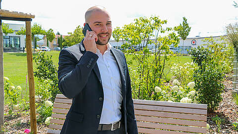 Ein Mann im dunklen Anzug steht im Freien und telefoniert mit seinem Smartphone. Er lächelt und steht neben einer Holzbank mit Grünflächen und einem Gebäude im Hintergrund.