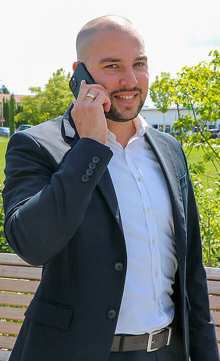 Vom Quereinsteiger zum top IT-Vertriebler Ein Mann im dunklen Anzug steht im Freien und telefoniert mit seinem Smartphone. Er lächelt und steht neben einer Holzbank mit Grünflächen und einem Gebäude im Hintergrund.