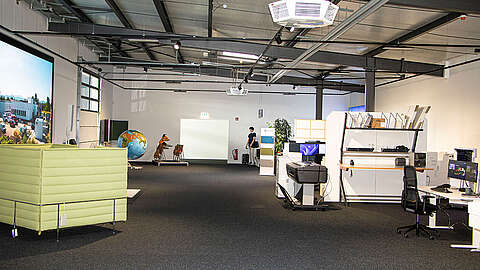 Ein modernes, geräumiges Büro mit Arbeitsplätzen, Computern, einem großen Globus, einem grünen Sofa und Regalen. Der Raum ist gut beleuchtet, hat freiliegende Decken und eine Person steht an der Rückwand.