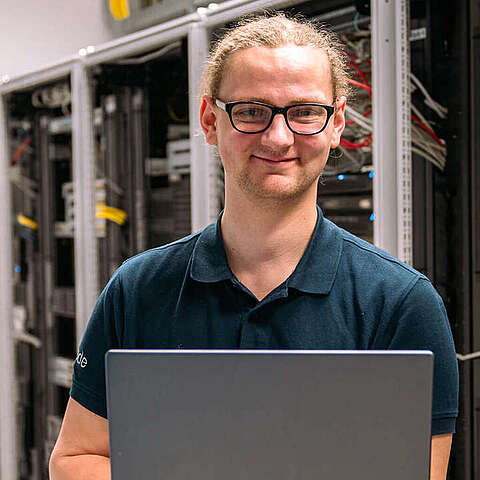 Auszubildender zum Fachinformatiker Systemintegration (m/w/d) steht vor einem Serverschrank und hält ein Laptop in der Hand