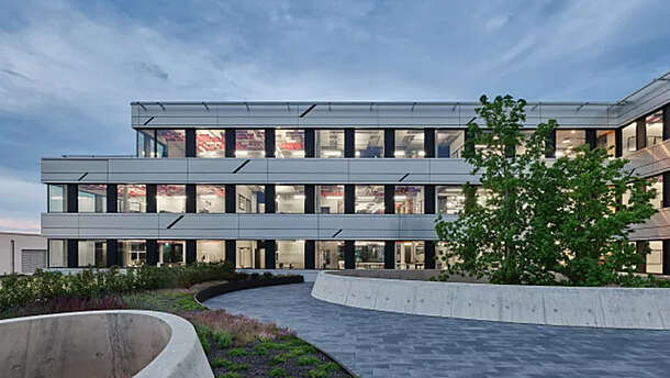 Modernes dreistöckiges Bürogebäude mit großen Fenstern, beleuchtetem Innenraum und einer Außenanlage mit Bäumen, Sträuchern und geschwungenen Betonwegen unter einem teilweise bewölkten Himmel in der Abenddämmerung.