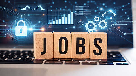 Auf Holz gedruckte Buchstaben, die das Wort "JOBS" ergeben, stehen auf der Tastatur eines Notebooks. Auf dem Bildschirm sind Symbole wie ein Schloss, Zahnräder oder ein Säulendiagramm zu sehen.