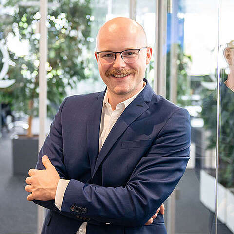 Führungsposition in der IT Ein lächelnder Mann mit Brille und Schnurrbart steht selbstbewusst mit verschränkten Armen in einem Büro. Er trägt einen blauen Anzug und ein weißes Hemd. Im Hintergrund sind Glaswände, Bürostühle und Pflanzen zu sehen.