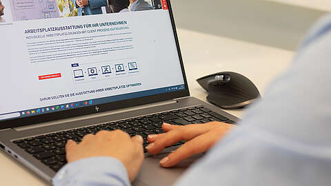 Eine Person tippt auf einem HP-Laptop und ruft eine Website über Büroausstattung auf. Neben dem Laptop liegt auf einem weißen Schreibtisch eine kabellose Maus. Auf der Website sind Symbole und Text auf Deutsch zu sehen.
