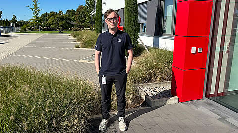 Eine Person, die eine Brille, ein marineblaues Polohemd, eine schwarze Hose und weiße Schuhe trägt, steht an einem sonnigen Tag vor einem modernen Gebäude mit roten und gläsernen Paneelen, umgeben von Grünflächen.