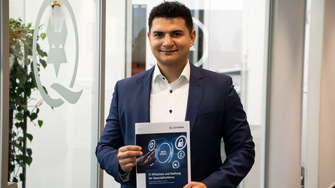 Ein Mann in einem marineblauen Anzug steht in einem Büro, lächelt und hält ein Dokument mit dem Titel "IT Pflichten und Haftung für Geschäftsführer" in der Hand. Im Hintergrund ist eine Milchglasscheibe mit einem Logo zu sehen.