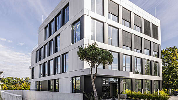 Ein modernes, vierstöckiges Bürogebäude mit großen Fenstern und weißer Vertäfelung, umgeben von ein paar Bäumen und Grünpflanzen unter einem blauen Himmel mit vereinzelten Wolken.