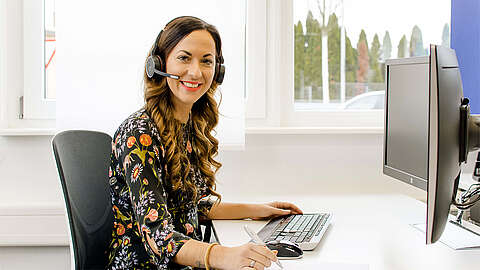 Frau sitzt an einem Schreibtisch mit PC und Tastatur, hat ein Headset auf und lächelt