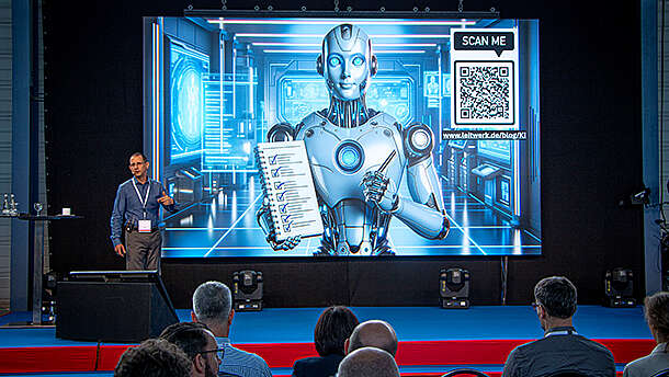 Ein Redner hält einen Vortrag vor einem Publikum, während hinter ihm auf einem großen Bildschirm ein humanoider Roboter zu sehen ist, der ein Dokument hält. Auf dem Bildschirm sind auch ein QR-Code und futuristische blaue Bilder zu sehen.