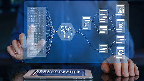 Eine Person interagiert mit einer transparenten digitalen Schnittstelle, die künstliche Intelligenz, Datenflussdiagramme und Grafiken anzeigt, während auf dem Tisch darunter ein Tablet mit einem Balkendiagramm liegt.