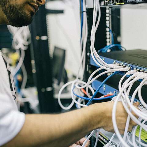 Eine Person in weißem Hemd verbindet oder organisiert mehrere Netzwerkkabel mit blauen Netzwerk-Switches, die auf einem Server-Rack montiert sind. Im Hintergrund sind weitere elektronische Geräte und Kabel zu sehen.