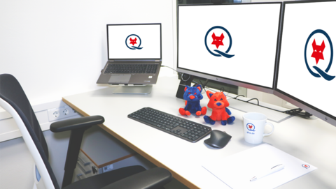 Höhenverstellbarer, weißer Schreibtisch mit Laptop und zwei großen Bildschirmen, auf denen das Q-FOX® Logo zu sehen ist. Vor den Bildschirmen sitzen ein blauer und ein roter Q-FOX®, außerdem sind auf dem Schreibtisch eine Q-FOX® Tasse, ein Block mit Kugelschreiber sowie eine Tastatur und Maus zu sehen.