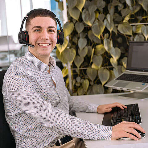 Auszubildender zum Kaufmann für IT-System-Management (m/w/d) sitzt am Schreibtisch, hat ein Headset auf und hält die Maus in der rechten Hand