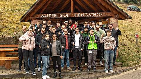Eine Gruppe von Menschen posiert und lächelt vor einem Holzgebäude mit dem Schild "Waldpension Hengsthof". Die meisten zeigen den Daumen nach oben, und die Einstellung scheint im Freien in einer ländlichen Gegend zu sein.