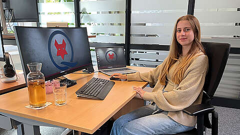 Eine Person mit langen blonden Haaren sitzt an einem modernen, höhenverstellbaren Schreibtisch mit einem großen Monitor und einem Laptop, die beide das Logo der Q-FOX® Gruppe zeigen. Auf dem Schreibtisch aus Holz stehen eine Karaffe Apfelschorle und ein Glas. Des Weiteren ist eine Tastatur sowie eine Maus zu sehen.