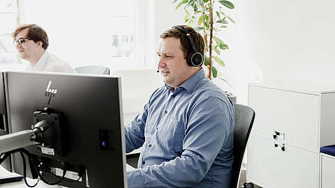 Zwei Männer arbeiten an Schreibtischen in einem hellen Büro. Der Mann im Vordergrund trägt ein blaues Hemd und ein Headset und schaut auf seinen Computerbildschirm. Im Hintergrund sind ein weiterer Mann, eine Pflanze und weiße Schränke zu sehen.