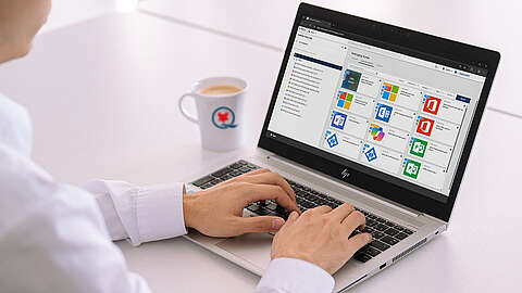 Eine Person tippt auf einem HP-Laptop, der einen Bildschirm für die Softwareverwaltung oder den App-Store anzeigt; eine Tasse mit einem blau-roten Logo und Kaffee steht in der Nähe auf einem weißen Schreibtisch.