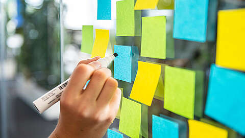Eine Person schreibt auf einen blauen Haftzettel, der an einer Glaswand befestigt ist, umgeben von anderen bunten Haftzetteln in Grün, Gelb und Blau, was auf Brainstorming- oder Planungsaktivitäten hindeutet.