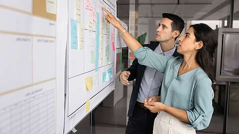 Zwei Kollegen stehen in einem modernen Büro vor einem Whiteboard mit Diagrammen und bunten Haftnotizen, diskutieren und zeigen auf die Tafel.