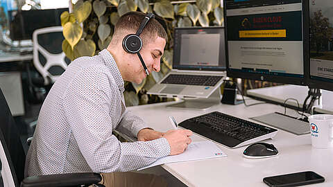 Moderne Arbeitsmittel: Warum sie für eine gute IT-Ausbildung entscheidend sind Eine junge Person mit Headset sitzt an einem Schreibtisch und schreibt auf Papier. Mehrere Monitore und ein Laptop stehen auf dem Schreibtisch, dazu eine Tasse und eine Computermaus. Im Hintergrund ist eine Blattpflanze zu sehen.
