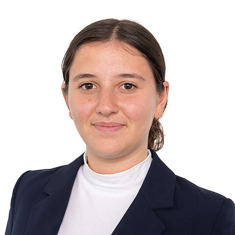 Eine junge Frau mit nach hinten gebundenen braunen Haaren, die einen marineblauen Blazer und einen weißen Rollkragenpullover trägt, lächelt leicht vor einem schlichten weißen Hintergrund.