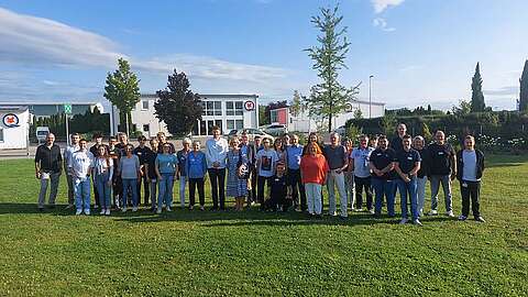 Eine große Gruppe von Menschen steht in Reihen auf einer Wiese im Freien und lächelt in die Kamera, während im Hintergrund moderne Gebäude, Bäume und ein blauer Himmel zu sehen sind.