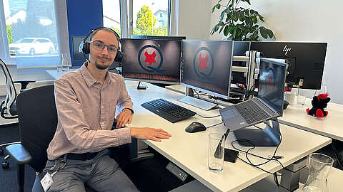 Eine Person, die eine Brille und ein Headset trägt, sitzt an einem modernen Schreibtisch mit drei Monitoren und einem Laptop, die alle das Logo der Q-FOX® Gruppe zeigen. Auf dem weißen Schreibtisch stehen ein Glas Wasser, Büroutensilien und ein kleiner Plüschfuchs.