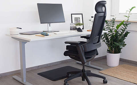 Ergonomischer schwarzer Bürostuhl steht vor einem weißen höhenverstellbaren Schreibtisch mit einem Laptop und einem weiteren Monitor. Auf dem Schreibtisch befinden sich weitere Gegenstände wie Tastatur, Notizbuch und Mäppchen, Trinken, eine Pflanze sowie zwei Bilder. Der Schreibtisch steht neben einem Fenster und einer Topfpflanze.