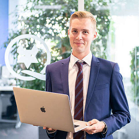 Karriere im IT-Vertrieb Eine Person in blauem Anzug und gestreifter Krawatte hält einen geöffneten Laptop in einer Büroumgebung. Im Hintergrund sind eine Glaswand, eine große Pflanze und Stühle um einen Tisch zu sehen, was auf einen modernen Arbeitsplatz hindeutet.