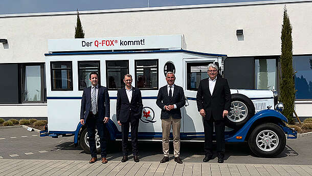 Vier Männer in Anzügen stehen vor einem weiß-blauen Oldtimer-Fahrzeug, auf dessen Dach ein Schild mit der Aufschrift "Der Q-FOX® kommt!" angebracht ist. Im Hintergrund sind ein modernes Gebäude und ein klarer Himmel zu sehen.