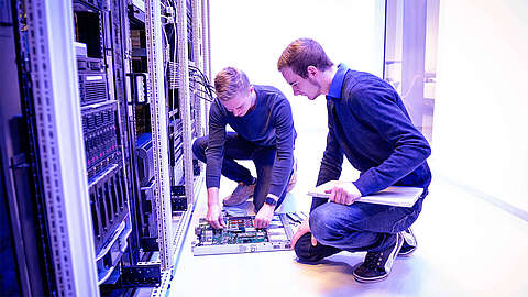 Zwei Mitarbeiter, die vor einem Serverschrank knien und an der Technik arbeiten