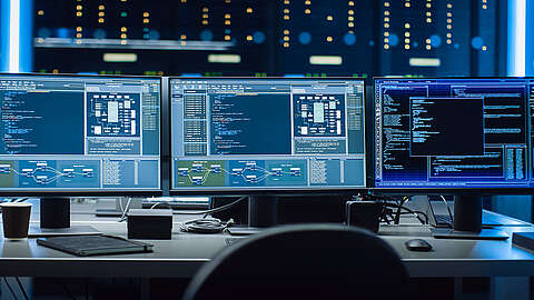 Drei Computermonitore auf einem Schreibtisch zeigen komplexe Codes, Diagramme und Systemdiagramme in einem schwach beleuchteten Kontrollraum oder Rechenzentrum an, wobei die Serverschränke im unscharfen Hintergrund zu sehen sind.