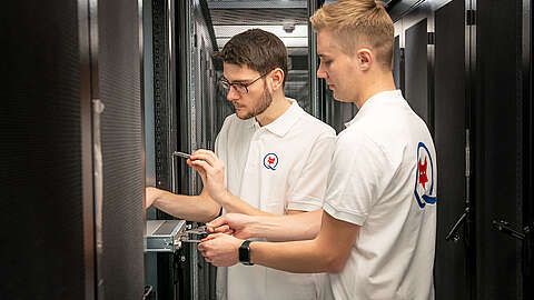 Zwei Männer in weißen Polohemden mit Logo arbeiten gemeinsam an Geräten in einem Serverraum und justieren und überprüfen Hardwarekomponenten in einem Netzwerkschrank.