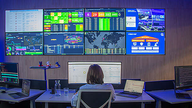 Eine Person sitzt an einem Schreibtisch mit drei Monitoren und blickt auf eine Wand mit großen Bildschirmen, auf denen Daten, Diagramme, Karten und Informationen zur Cybersicherheit in einem modernen Kontrollraum angezeigt werden.