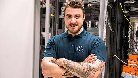 Praxisorientierung: Der Schlüssel zu einer erfolgreichen IT-Ausbildung Eine junge Person mit Tattoos und Bart steht selbstbewusst mit verschränkten Armen in einem Serverraum und trägt ein blaues Poloshirt. Im Hintergrund sind Racks mit Servern und Netzwerkgeräten zu sehen.