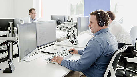 Individuelle Softwareentwicklung Eine Person mit Kopfhörern arbeitet an einem Schreibtisch mit zwei Monitoren. Mehrere andere sitzen an Schreibtischen in der Nähe und benutzen ebenfalls Computer. Das Büro ist hell und modern gestaltet, mit weißen Möbeln und natürlichem Licht.