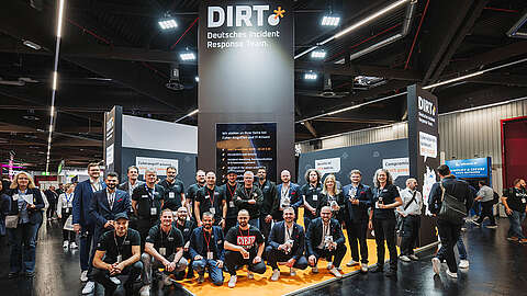 Eine Gruppe von Personen posiert gemeinsam vor einem großen Stand mit der Aufschrift "DIRT Deutsches Incident Response Team" auf einem Kongress. Die Umgebung ist hell und geschäftig, mit anderen Ständen und Teilnehmern im Hintergrund.