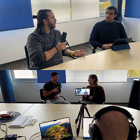 Zwei Männer sitzen an einem Tisch und führen ein Gespräch, einer hält ein Mikrofon in der Hand. In einer anderen Ansicht sind eine Kamera und ein Laptop aufgestellt, um das Gespräch aufzuzeichnen, wobei im Vordergrund eine Person mit Kopfhörern zu sehen ist.