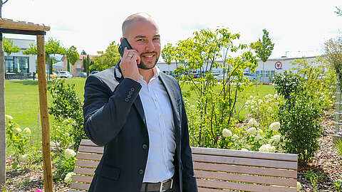 Ein Mann im Anzug steht im Freien in der Nähe einer Holzbank, lächelt und spricht mit einem Mobiltelefon, während im Hintergrund grüne Pflanzen und Gebäude zu sehen sind.