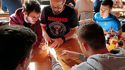 Eine Gruppe sitzt um einen Holztisch und konzentriert sich darauf, eine Konstruktion aus Holzstäbchen zu bauen. Auf dem Tisch stehen Limonadenflaschen. Sonnenlicht scheint auf die Hände.