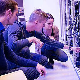 Eine knieende Person zeigt zwei anderen Personen etwas an einem Serverschrank