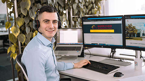 Q-FOX® Mitarbeiter sitzt an einem Schreibtisch mit PC-Monitoren und hat ein Headset auf