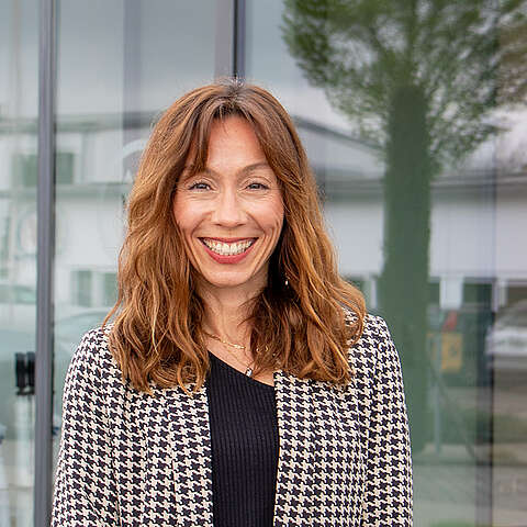 Eine Frau mit langen braunen Haaren lächelt, während sie im Freien vor einem belaubten Baum steht. Sie trägt ein schwarzes Hemd und einen schwarz-weißen Blazer mit Hahnentrittmuster. Der Hintergrund besteht aus grünem Gras und unscharfem Laub.