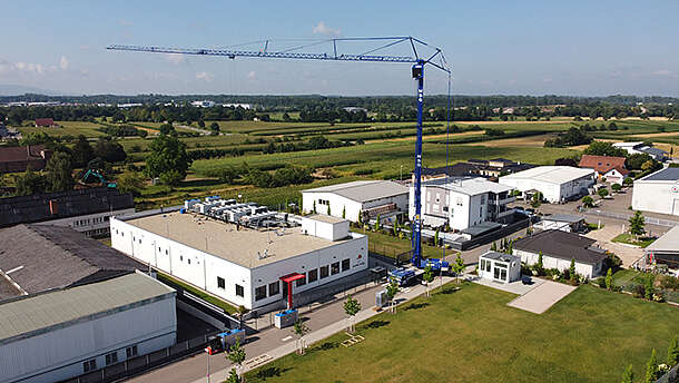 Ein großer blauer Baukran steht in der Nähe eines weißen Industriegebäudes, umgeben von grünen Feldern, anderen Gebäuden und Straßen, unter einem teilweise bewölkten Himmel.