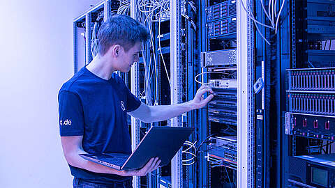 Ein Techniker mit einem Laptop passt Kabel an einem Serverschrank in einem Rechenzentrum an, umgeben von blauer Beleuchtung und verschiedenen Netzwerkgeräten.