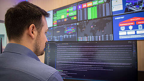 Ein Mann in einem blauen Hemd arbeitet an einem großen Computermonitor, auf dem Codes angezeigt werden, während im Hintergrund mehrere bunte Daten- und Sicherheitstafeln auf Bildschirmen an der Wand angebracht sind.